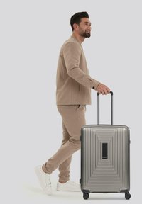 Uomo in abbigliamento beige che cammina e trascina una valigia rigida argentata con quattro ruote e un manico telescopico.