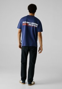 Tamsiai mėlynos medvilnės marškinėliai su užrašu „RED BULL RACING RRR GIVES YOU WINGS“ ryškiomis baltomis ir raudonomis raidėmis ant nugaros. Laisvas siluetas ir trumpomis rankovėmis.