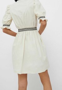 Cremefarvet kjole med korte pufærmer, samlet talje og sorte broderede detaljer ved taljen og ærmerne. Frit skørt design.