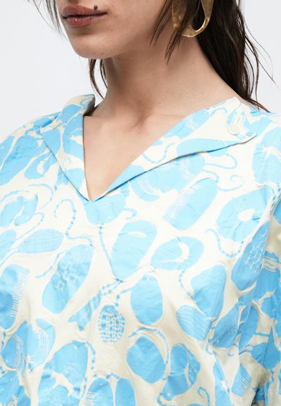 Γυναίκα που φορά ένα λευκό πουκάμισο με γιακά και ανοιχτό μπλε αφηρημένο σχέδιο, ένα χρυσό σκουλαρίκι και μια ελιά κοντά στο στόμα της.