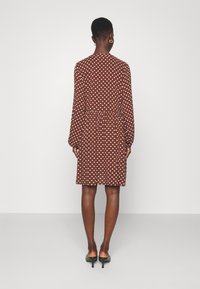 Vestido marrón con lunares, de mangas largas, corte ajustado, cintura fruncida y largo hasta la rodilla. Textura suave y tela blanda, combinado con zapatos de tacón.