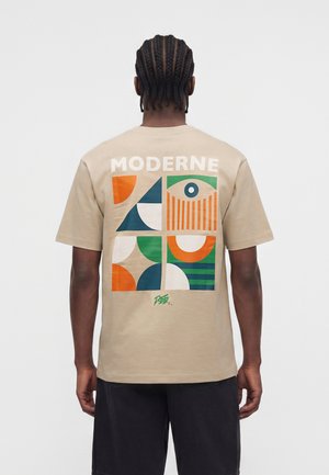 Hombre con el pelo trenzado que lleva una camiseta beige con el texto "MODERNE" y formas geométricas abstractas de colores en la espalda, combinada con pantalones cortos oscuros.
