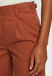 Pantalon couleur rouille à texture lisse, doté d'un design plissé et d'une taille ajustée. Des coutures visibles et des passants de ceinture ajoutent du détail.