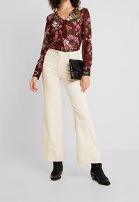 Kobieta w kremowych, szerokich spodniach, czarnych botkach za kostkę, bordowej, kwiecistej bluzce z długim rękawem i małej, czarnej torebce na ramię.