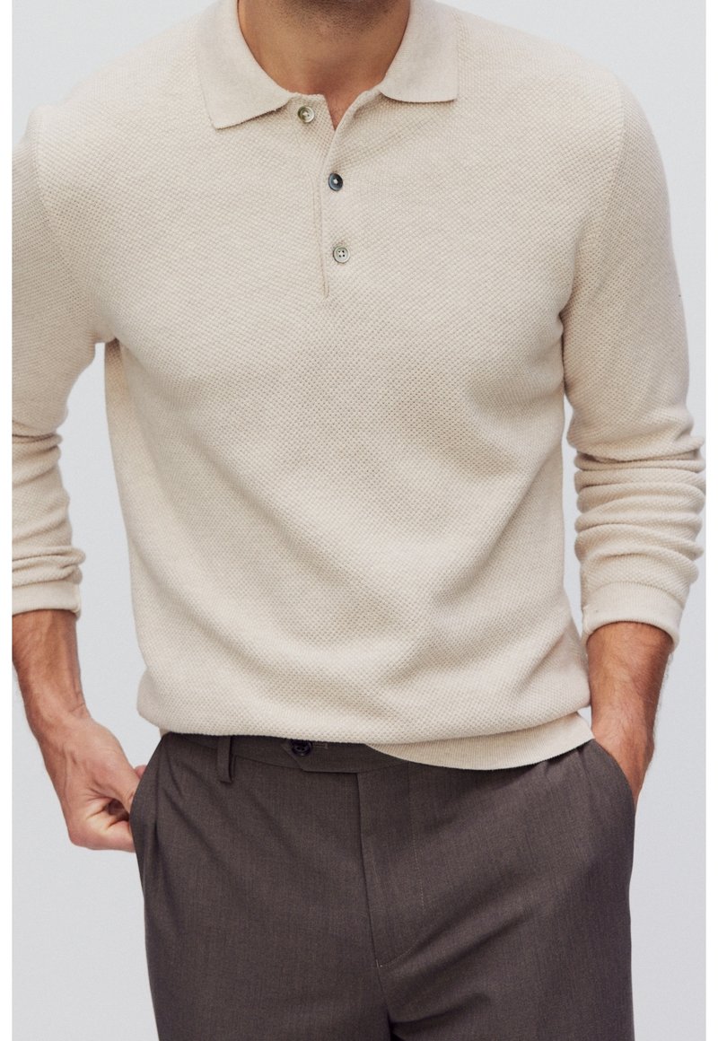 Homme portant un polo texturé beige à manches longues avec trois boutons et un pantalon gris foncé, les mains dans les poches, cadré au niveau du cou et des hanches.