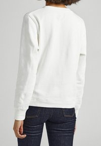 Felpa bianca in cotone con maniche lunghe, polsini a coste e scollo rotondo; abbinata a jeans in denim scuro. Nessun logo o motivo visibile.