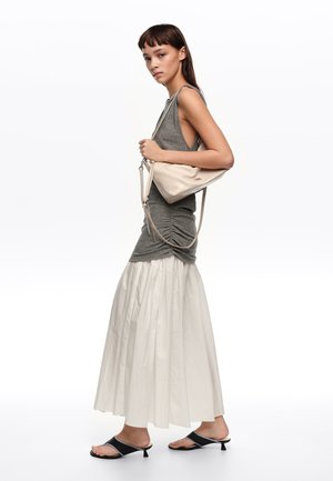 Mujer con camiseta sin mangas gris, falda larga blanca, sandalias negras de tacón bajo y bolso beige de hombro, de pie de perfil.