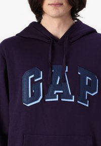 Felpa con cappuccio viola caratterizzata da un grande logo "GAP" in feltro sovrapposto, con finitura testurizzata e accenti azzurri a contrasto.
