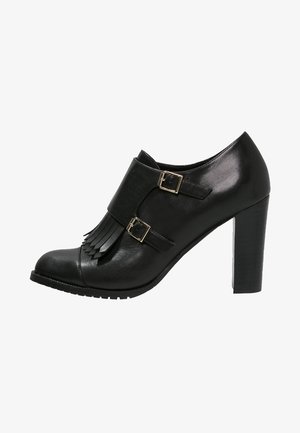 Bottine noire à talons hauts, avec un bout arrondi, des boucles dorées et des franges sur le devant. En cuir lisse avec une semelle texturée.