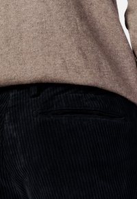 Brauner Strickpullover über schwarzer Cordhose mit vertikaler Rippoptik; die Hose hat eine dezente Gesäßtasche.