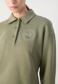Femme portant un polo à manches longues vert olive avec trois boutons et un logo Lacoste brodé sur le côté gauche de la poitrine, sur un fond uni.