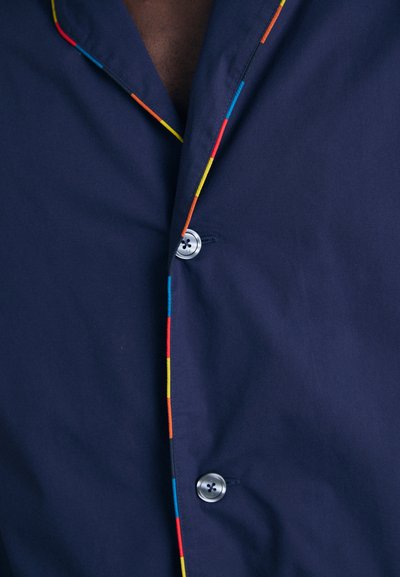Chemise bleu marine avec une texture lisse, ornée de passepoils colorés le long du col et d'une fermeture à boutons gris au niveau de la poitrine.