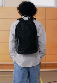 Fekete hátizsák letisztult dizájnnal, tartós szövetből készült. Szögletes elülső panellel, cipzáras zsebekkel és a tetején logó díszítéssel rendelkezik.