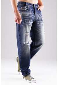 Jeans aus Denim mit einem verblassten blauen Farbverlauf, verschlissenen Mustern und mehreren Rissen. Kombiniert mit olivgrünen Sneakers mit weißen Sohlen.