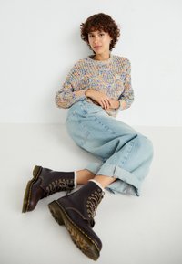 Un pull en maille multicolore avec un motif texturé, un jean bleu clair, et des bottes en cuir marron à lacets avec des bordures jaunes et des semelles robustes.