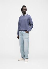 Sweatshirt azul claro com decote em V e logótipo bordado a amarelo, combinado com jeans de lavagem clara de corte largo e calçado bege.