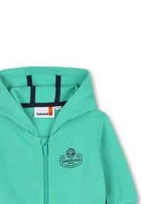 Sweat à capuche vert zippé avec capuche, logo Timberland et accents bleu marine. Fabriqué en tissu doux et texturé avec finitions côtelées.