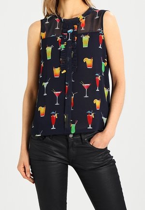 Vrouw draagt mouwloze marineblauwe blouse met patroon van kleurrijke cocktails, gecombineerd met zwarte leren broek, hand in de zak.