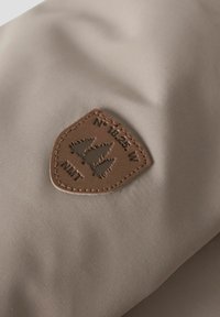 Pruun nahkpatch, millel on kolm männit树d ja kiri "N° 18.25 W NMT", õmmeldud beežile kangale.