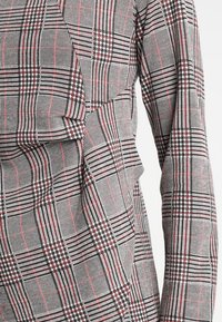 Chemise à carreaux grise avec des accents noirs et rouges, réalisée en tissu texturé aux motifs variés, manches longues et coupe cintrée.