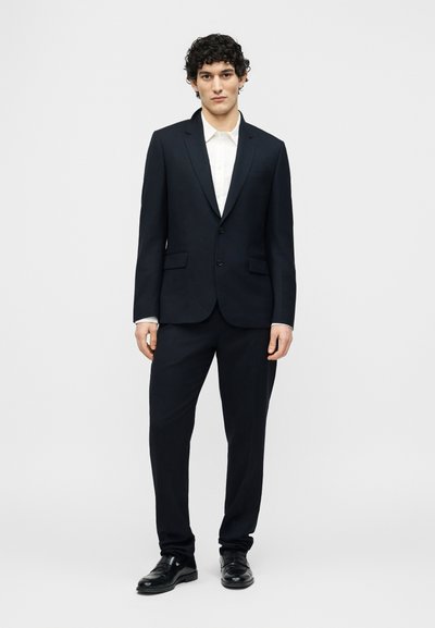 Homme aux cheveux bouclés portant un costume noir à deux boutons, une chemise blanche et des chaussures en cuir noires, debout devant un fond clair uni.