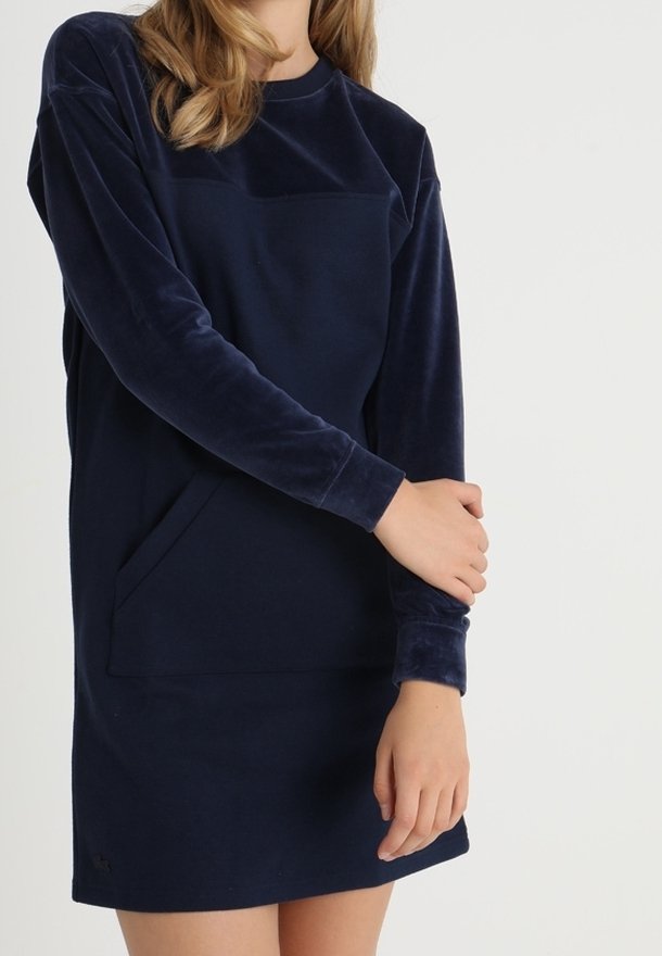 Femme portant une robe en velours bleu marine à manches longues avec une poche avant, posant avec un bras croisé sur l'autre.