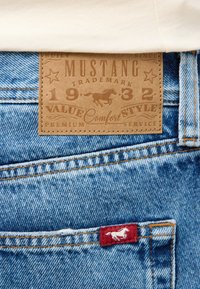 Blaue Denim-Jeans mit einem braunen Lederetikett, das geprägtem Text und einem Pferdedesign aufweist. Rotes Etikett an der Gesäßtasche mit weißem Pferdelogo.