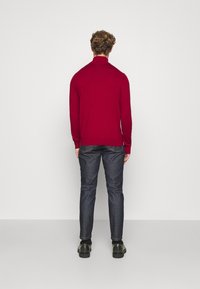 Punainen neulepaita, jossa on korkea kaulus, pitkät hihat ja sileä pinta, yhdistettynä tummansinisiin slim-fit-farkkuihin ja mustiin kenkiin.