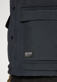 Mörkblå puffjacka med knäppning, framficka och synlig varumärkesetikett. Materialet verkar texturerat och isolerat.