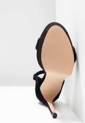 Schwarzer High-Heel-Schuh, der mit der Sohle nach oben an einer weißen Eckwand steht und eine glatte, beige Sohle mit strukturiertem Griffbereich zeigt.