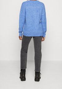 Person klädd i en blå strukturerad tröja, grå raka jeans och svarta kraftiga stövlar som står mot en enkel ljus bakgrund.