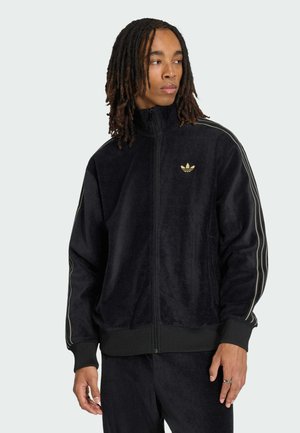 Giovane con dreadlocks alle spalle che indossa una giacca da ginnastica nera Adidas con logo e righe dorate, guarda verso destra.