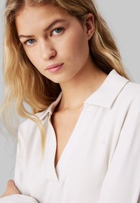 Biały, teksturowany sweter z dekoltem w serek oraz kołnierzykiem. Posiada subtelną markę po lewej stronie. Modelka ma falowane, blond włosy i neutralny makijaż.