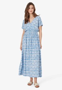 Maxikleid aus leicht blauem Stoff mit geometrischem Muster und kurzen Ärmeln. Elastischer Taillenbund und lockere Passform. Kombiniert mit hellen Sandalen.