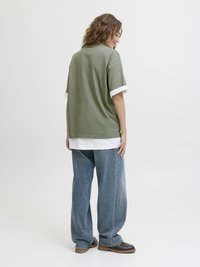 Olivenfarget oversized t-skjorte med hvite mansjett-detaljer, kombinert med løse blå denimjeans og mørkebrune sko, sett fra baksiden.