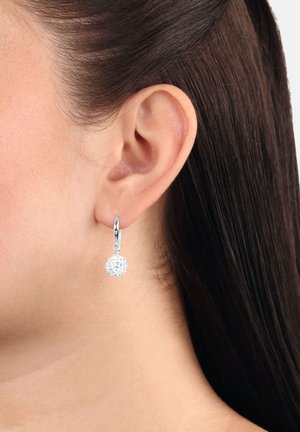 Orecchini a cerchio argentati con un pendente rotondo scintillante incastonato di pietre trasparenti, mostrati all'orecchio di un modello con capelli scuri e lisci.