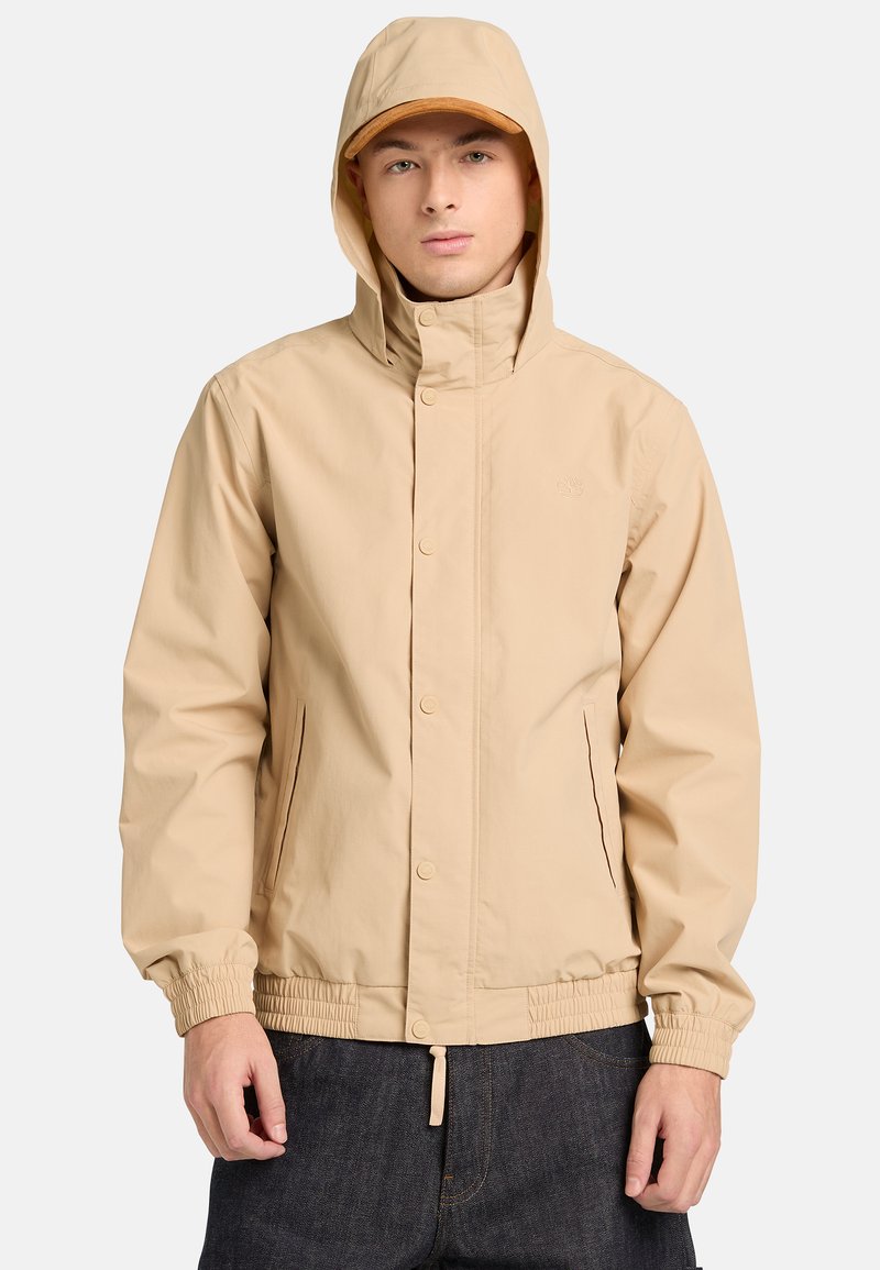 Joven con chaqueta beige con capucha, broches a presión y puños elásticos, combinada con pantalones de mezclilla oscuros.