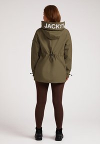 Oliwkowa wodoodporna kurtka z regulowanym pasem, dużym kapturem i białym napisem "JACK1T" na plecach. Posiada długie rękawy i regulowane mankiety.