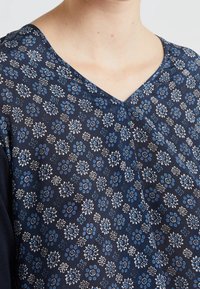 Tummansininen paita, jossa on V-pääntie ja vaaleansinisestä ja valkoisesta koostuva kukkakuosi. Pehmeää kangasta, jossa on teksturoitu pinta.