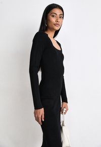 Black knitted dress with long sleeves, scoop neckline, and vertical ribbed texture. Model holds a small white handbag.