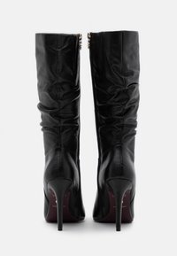 Botas de cuero negras hasta la rodilla con diseño fruncido, punta afilada y tacón de aguja de 4 pulgadas, con cremalleras laterales para el cierre.