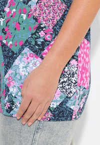 Barevný květinový patchwork top s různými vzory v růžové, zelené a tmavě modré barvě, vyrobený z měkké látky a s uvolněným střihem. Ruka odpočívá na oděvu.