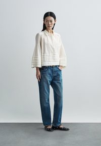 Valkoinen kauluksellinen pusero, jossa pystysuorat laskokset, yhdistettynä rennosti istuviin sinisiin farkkuihin ja mustiin litteisiin kenkiin, joissa on koristeellisia metalliyksityiskohtia.