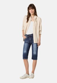 Chemise beige à boutons sur un débardeur blanc, portée avec un short en denim bleu foncé jusqu'aux genoux et des baskets blanches. Look détendu et décontracté.