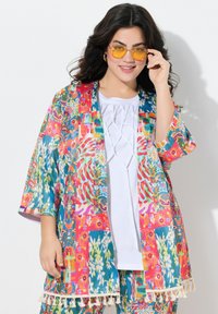 Kimono leggero e colorato con motivi floreali e astratti, caratterizzato da un orlo con frange e maniche ampie. Abbinato a una maglietta bianca.