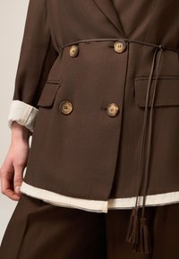 Bruine dubbelzijdige blazer met tan knoopaccenten en een strikceintuur. Voorzien van voorzakken en een contrasterende lichte zoom en manchetten.