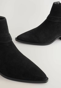 Botas de tobillo de ante negro con punta afilada, tacones bajos de bloque y una correa decorativa alrededor del tobillo para añadir detalle.