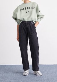 Ljusgrön oversized sweatshirt med "NIKE"-logotyp, i kombination med löst sittande svarta cargo-byxor med sidofickor och guld detaljer, bärs med silverfärgade sneakers.