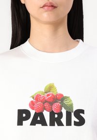 T-shirt en coton blanc avec un motif de framboises et de feuilles vertes au-dessus du mot "PARIS" en lettres noires en gras.