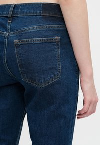 Zbliżenie dolnej części pleców i ramienia osoby noszącej ciemnoniebieskie dżinsy z widocznymi szwami i tylną kieszenią.